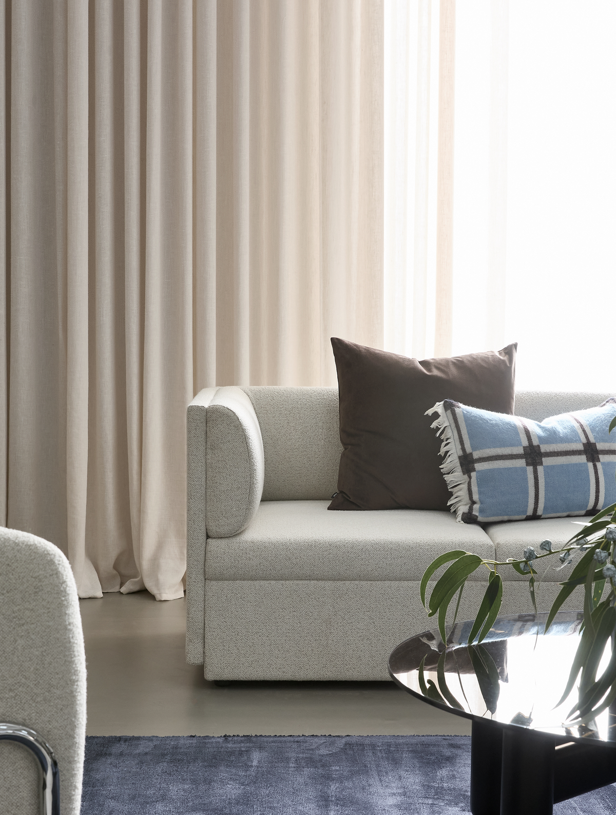 Ljus modern vardagsrumsdel med beige gardiner, ljus boucléssoffa med brun kudde och blå-rutigt kuddfodral, glas soffbord med gröna kvistar och blå matta.