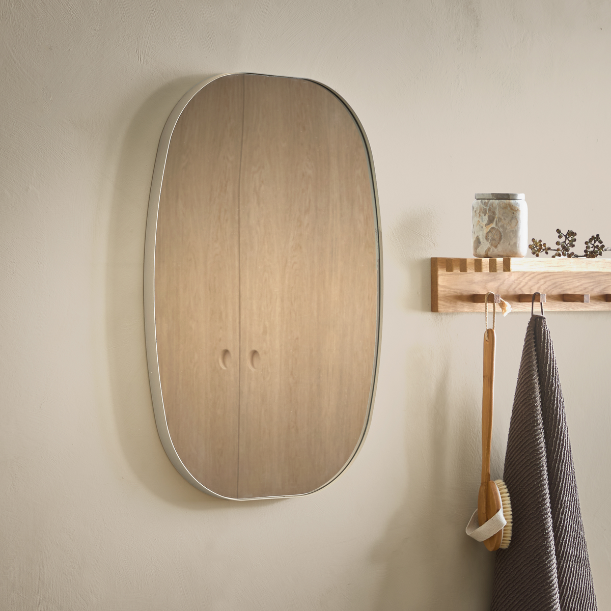 Organisk oval väggspegel med tunn metallram bredvid en trälist med krokar, burk, badborste och mörk handduk i ett beige badrum.