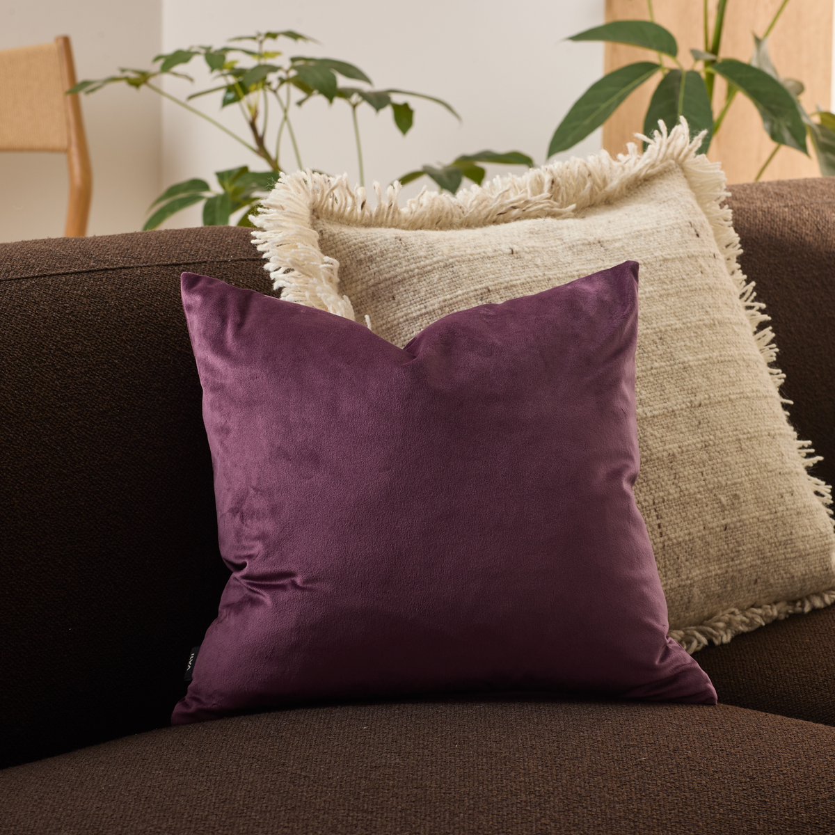 Plommonlila prydnadskudde på mörkbrun soffa framför beige kudde med fransar; växter i bakgrunden.