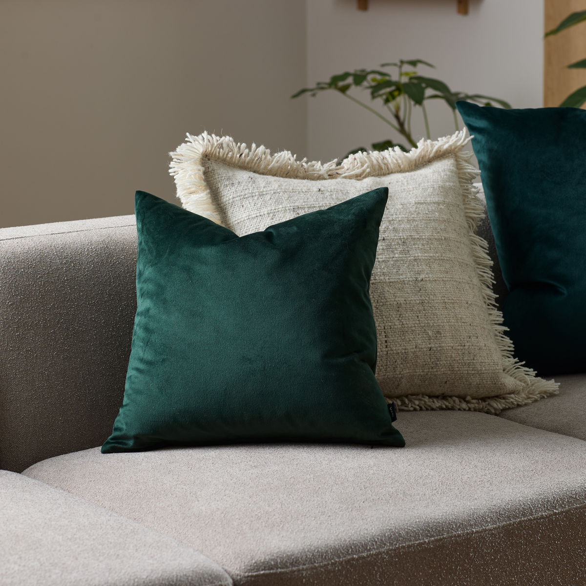 Mörkgrön sammetskudde och beige franskantad kudde på beige soffa i vardagsrum.
