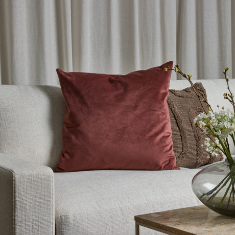 Ljus soffa med stor roströd sammetskudde och brun strukturkudde; glasvas med vita blommor på bord framför ljusa gardiner.