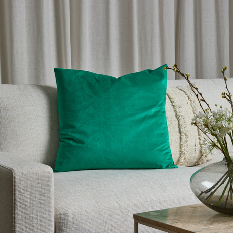 Grön prydnadskudde på ljus soffa med neutrala kuddar, beigea gardiner bakom och en glasvas med vita blommor på soffbordet.