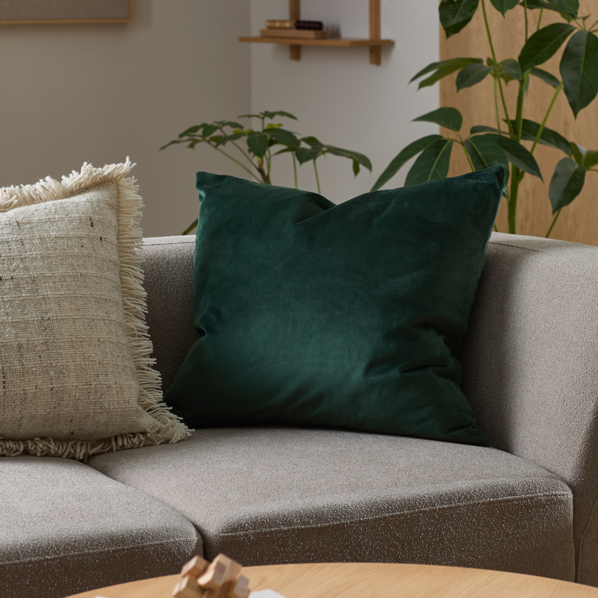 Grön sammetskudde på grå soffa i vardagsrum med beige franskudde och gröna växter.