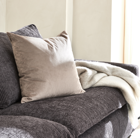 Beige sammetskudde på mörk ribbad soffa med mjuk vit pläd i dagsljus vid fönster.