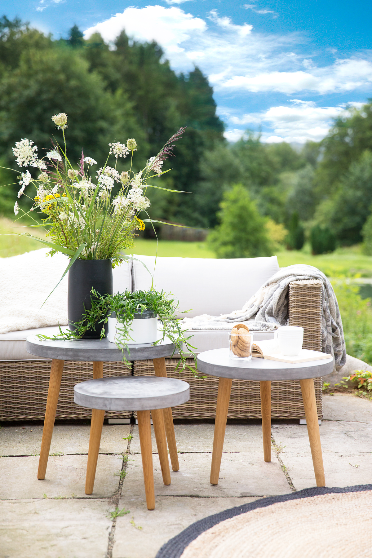 Somrig uteplats med rottingsoffa, tre små runda bord med betongskivor och träben, vilda blommor i svart vas, grön krukväxt, mjuk pläd, bok, kopp och glas med kakor mot grönskande träd och blå himmel.