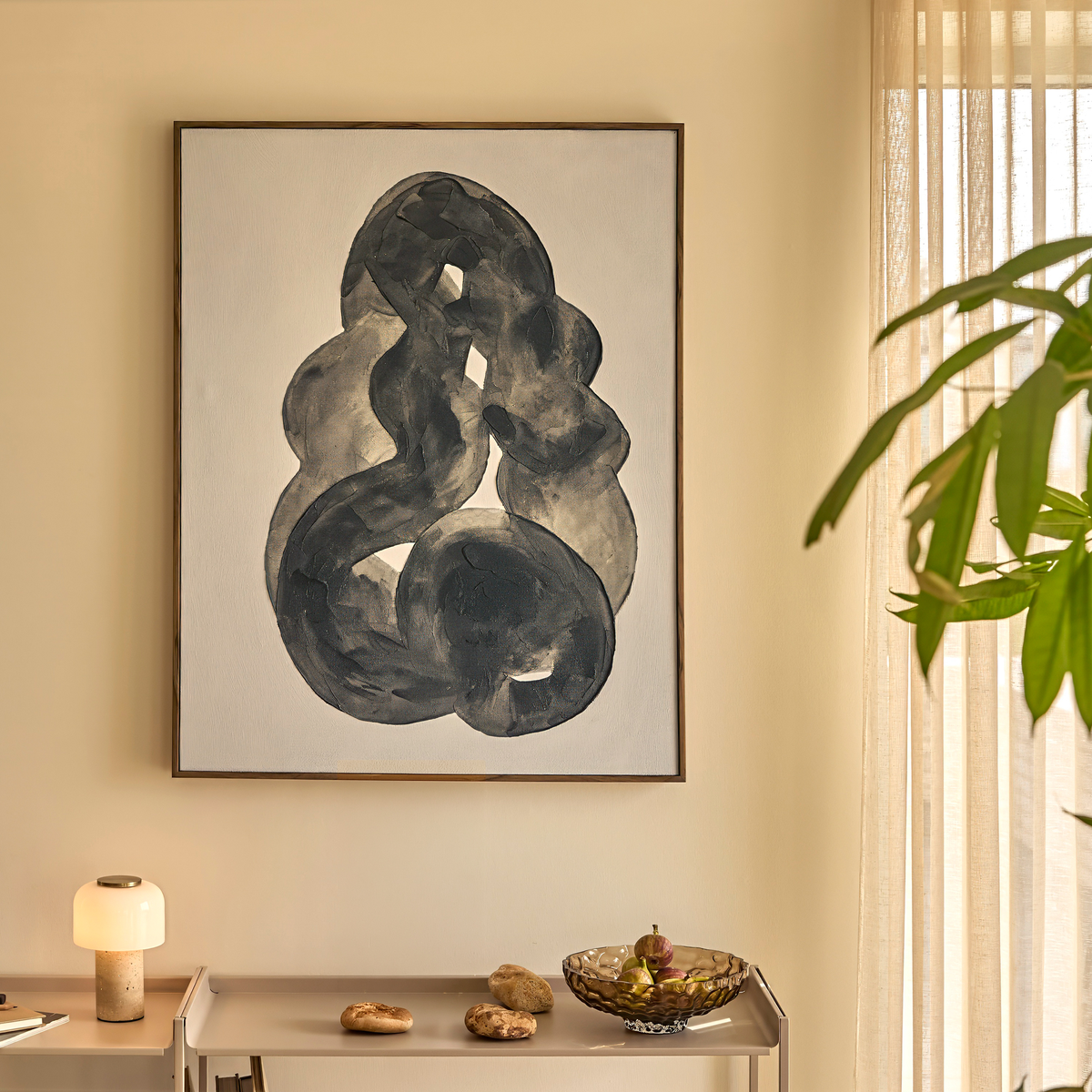 Abstrakt mörkgrå målning på beige vägg ovanför smal konsol med liten bordslampa, skål med frukt och några stenar; mjukt dagsljus genom ljusa gardiner och grön växt i kanten.
