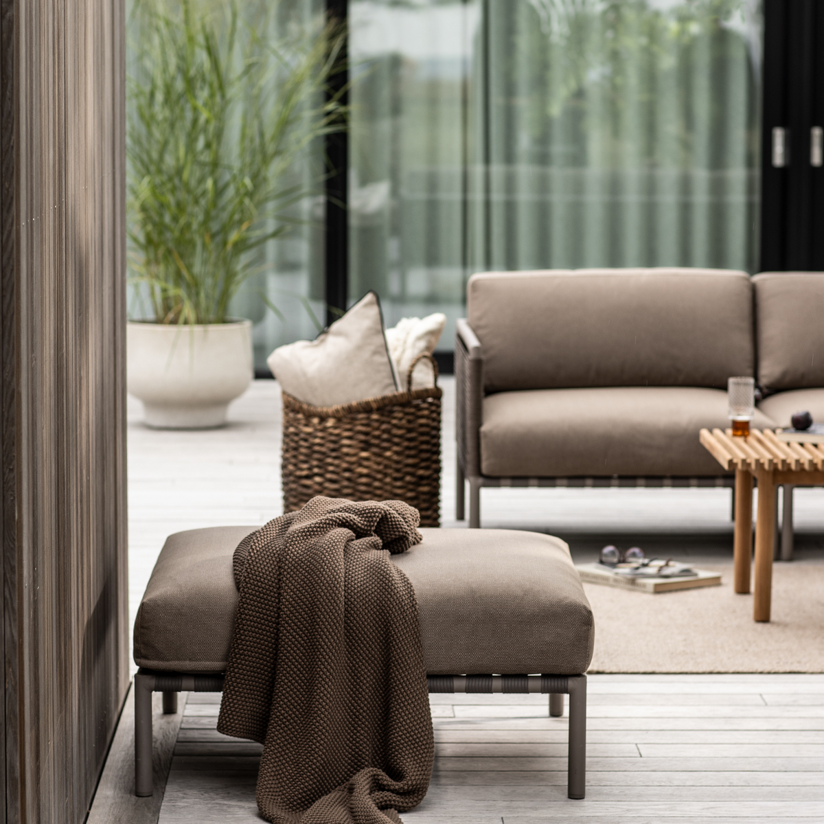 Uteplats med beige loungemöbler – en ottoman med brun stickad pläd i förgrunden, soffa och träbord, korg med kuddar samt grön växt i kruka.