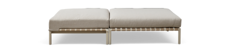 Modulär ottoman/bänk med två beige sittdynor på ljus metallram med vävd rembas, sedd rakt framifrån på mörk bakgrund.