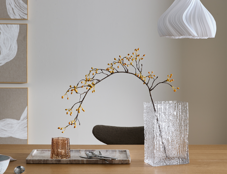 Rektangulär klar, texturerad glasvas på ett träbord med en böjd gren med gula bär; bredvid en rosa ljuslykta på en stenbricka, en vit veckad pendellampa ovanför och neutrala väggtavlor i bakgrunden.