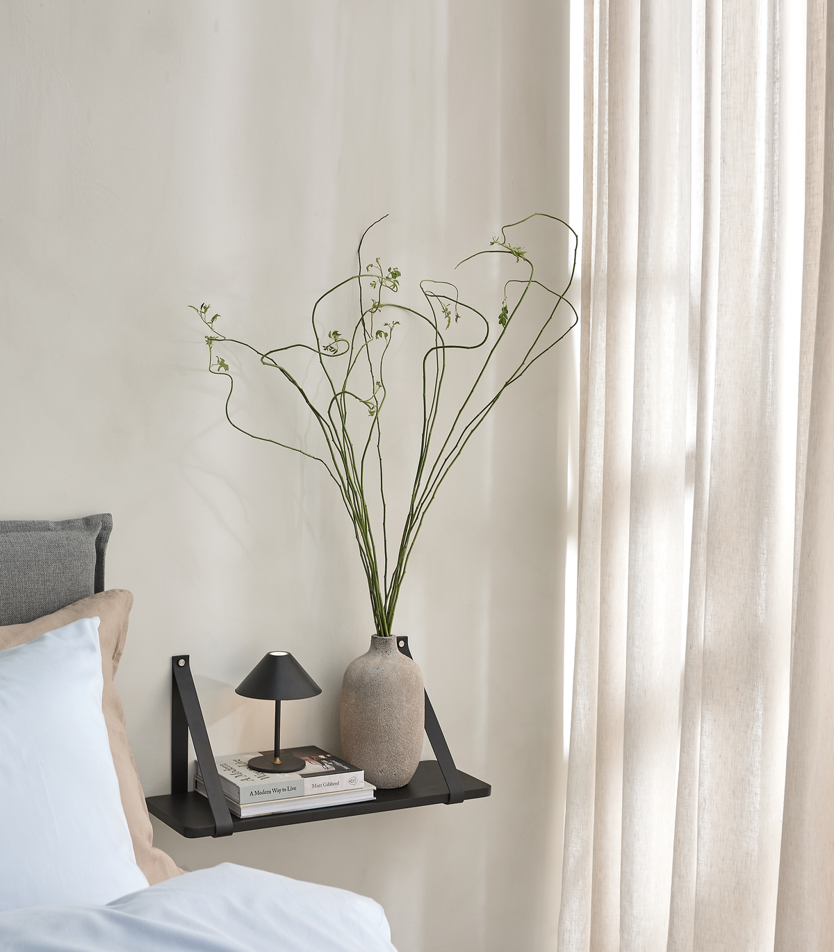 Ljust sovrum med svart vägghylla som sängbord, liten svart bordslampa, böcker och beige vas med höga gröna kvistar; linnegardiner i dagsljus.