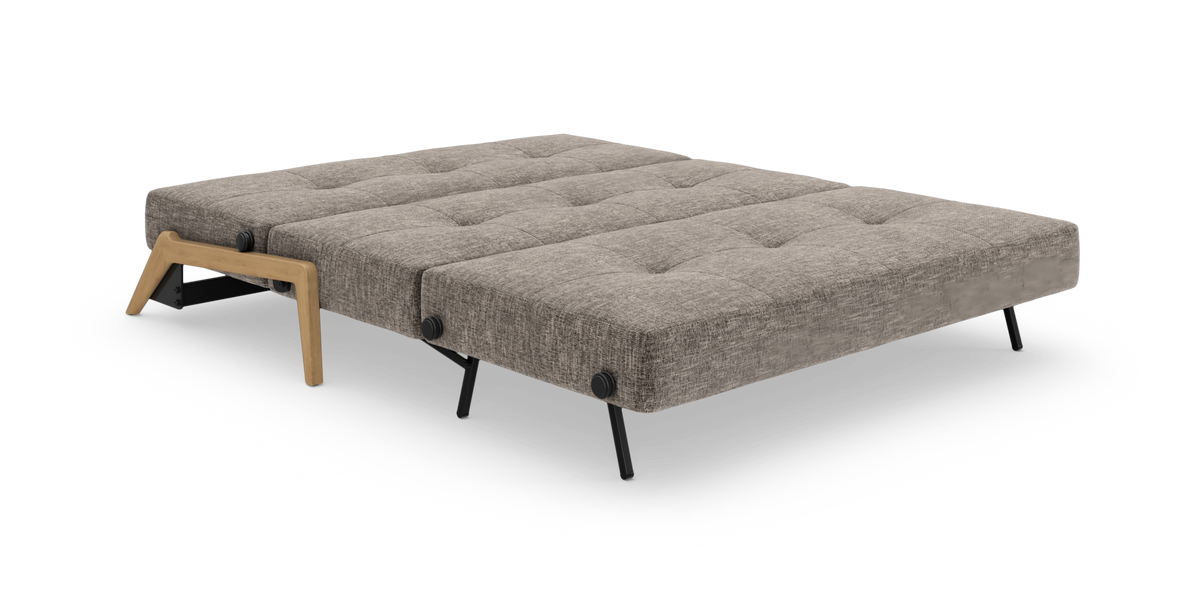 Uppfällbar bäddsoffa i helutfällt läge med grå‑beige tyg, svarta ben och trädetaĺj vid armstödet, frilagd mot vit bakgrund.