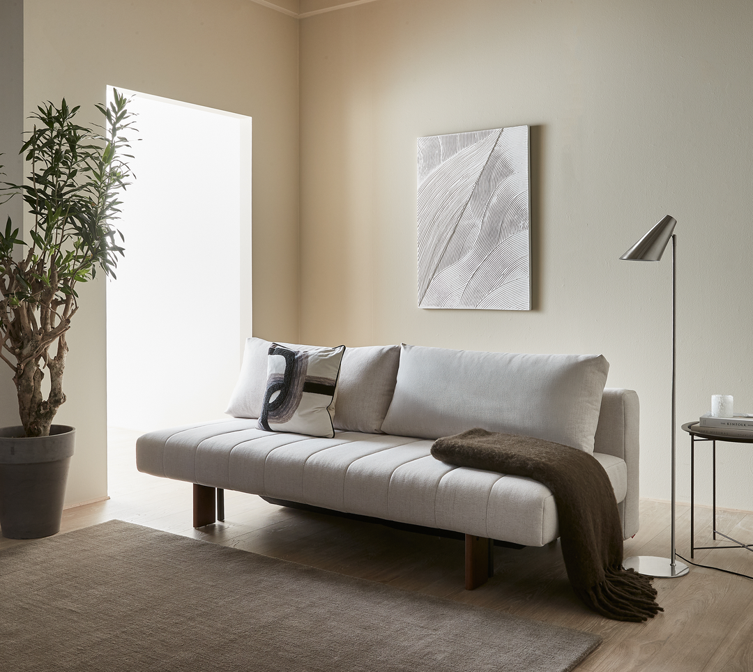 Ljus minimalistisk vardagsrumsscen med beige soffa med kuddar och brun pläd, golvlampa och sidobord med ljus och boken Kinfolk, väggkonst och stor växt vid ljust fönster.