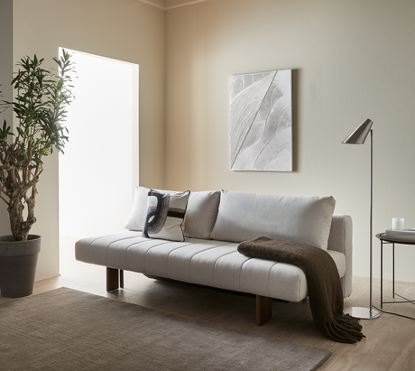 Ljus minimalistisk vardagsrumsscen med beige soffa med kuddar och brun pläd, golvlampa och sidobord med ljus och boken Kinfolk, väggkonst och stor växt vid ljust fönster.