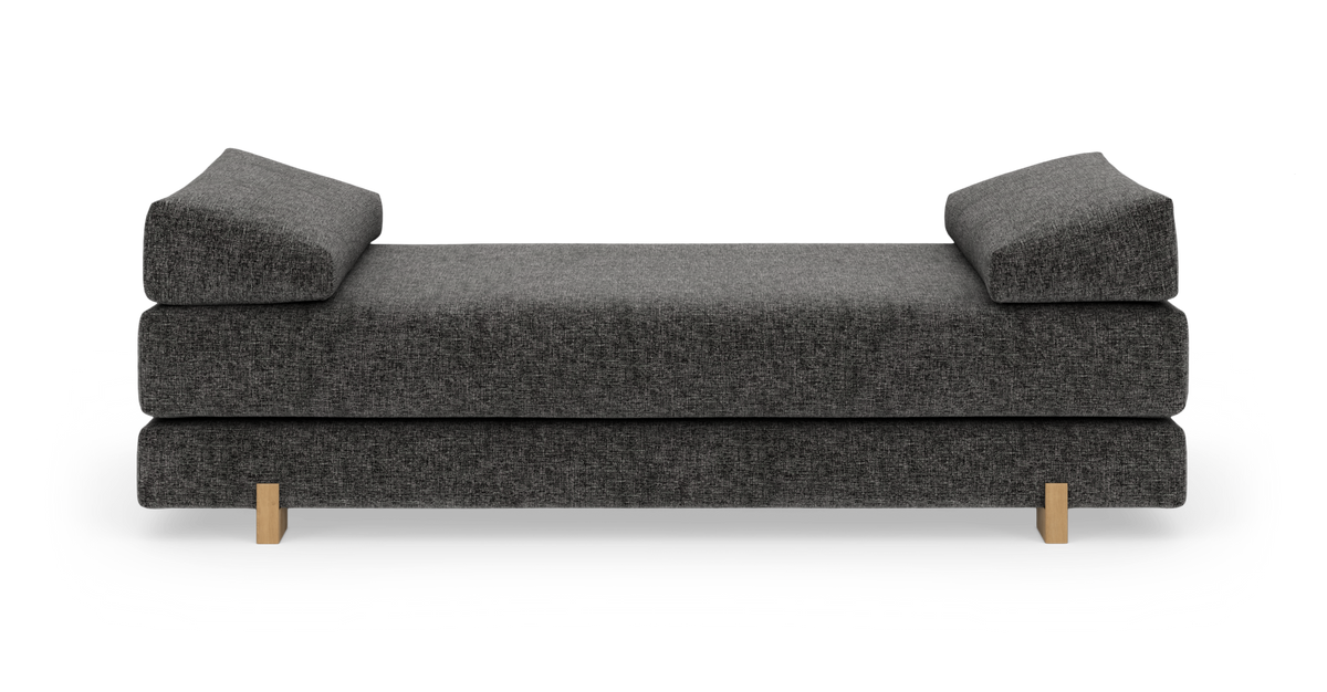 Mörkgrå daybed/soffa med två skrånande pudestöd och lyse träben, sedd framifrån på vit bakgrund.