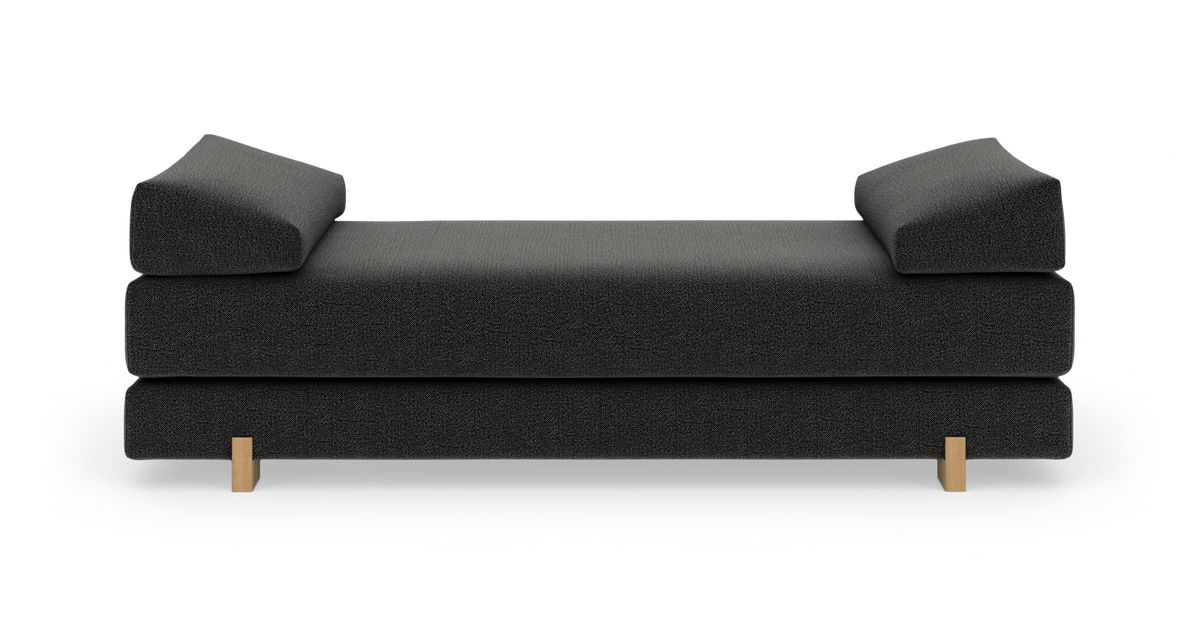 Mörkgrå dagbädd/soffa med två kilformade armstöd och ljusa träben, frilagd mot vit bakgrund i frontvy.