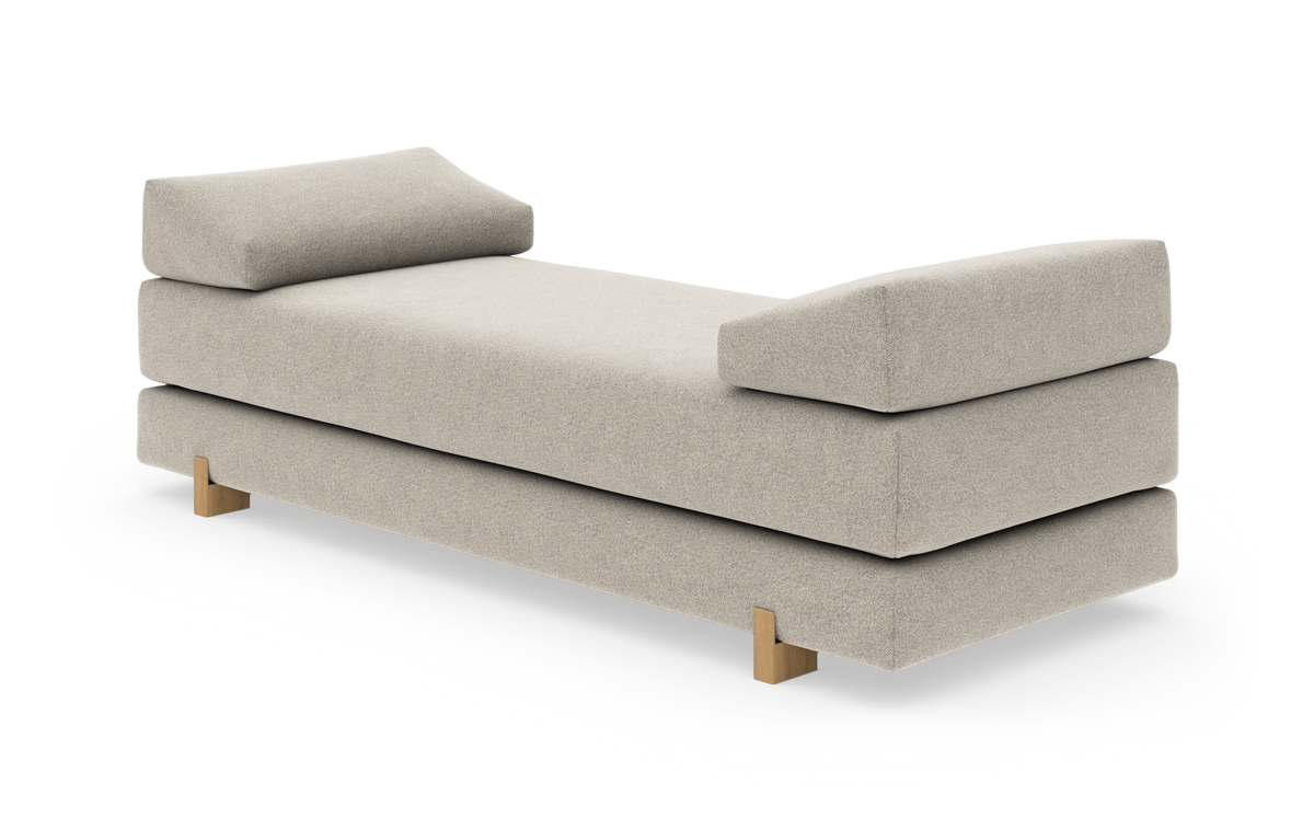 Beige daybed med två snedställda kuddade armstöd och träben, frilagd på vit bakgrund.