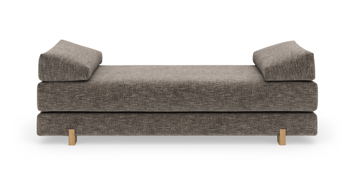 Grå stoppad daybed med två lutande kuddar och ljusa träben, fotad rakt fram mot vit bakgrund.