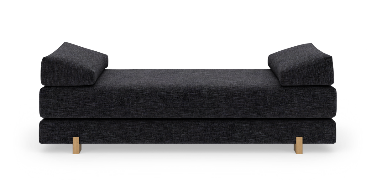 Mörkgrå dagbädd/soffa med två kilformade armkuddar och ljusa träben, frilagd på vit bakgrund.