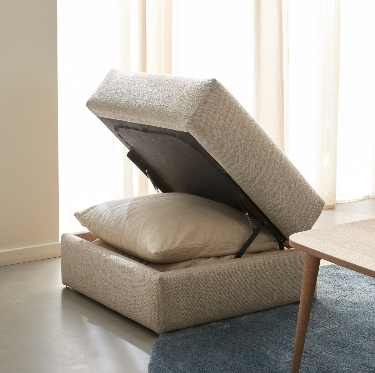 Beige förvaringspall med uppfällbart lock och kudde inuti i ett ljust vardagsrum.