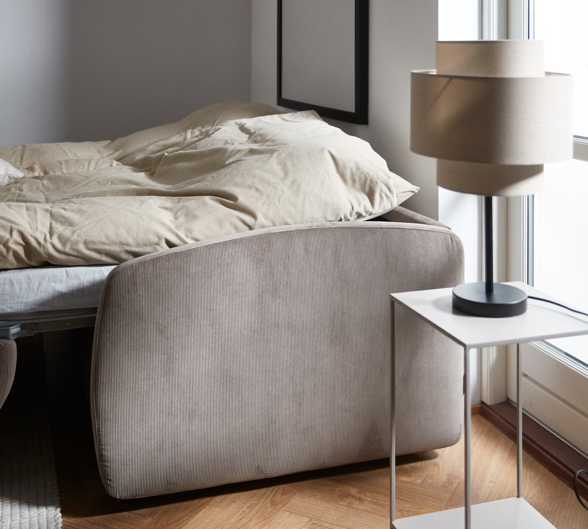 Ljust sovrum med beige sänggavel, bäddad säng och modern bordslampa på vitt sidobord vid fönster.
