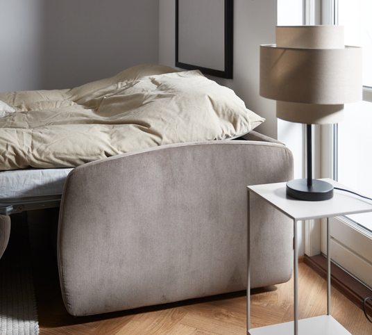 Ljust sovrum med beige sänggavel, bäddad säng och modern bordslampa på vitt sidobord vid fönster.