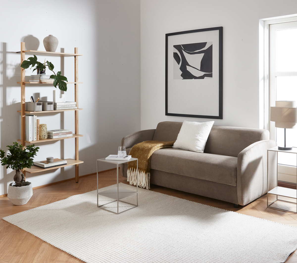 Ljust vardagsrum i skandinavisk stil med beige soffa, vit kudde och senapsgul pläd, litet metallbord, hylla med böcker och växter, stor ljus matta och abstrakt tavla vid ett fönster.