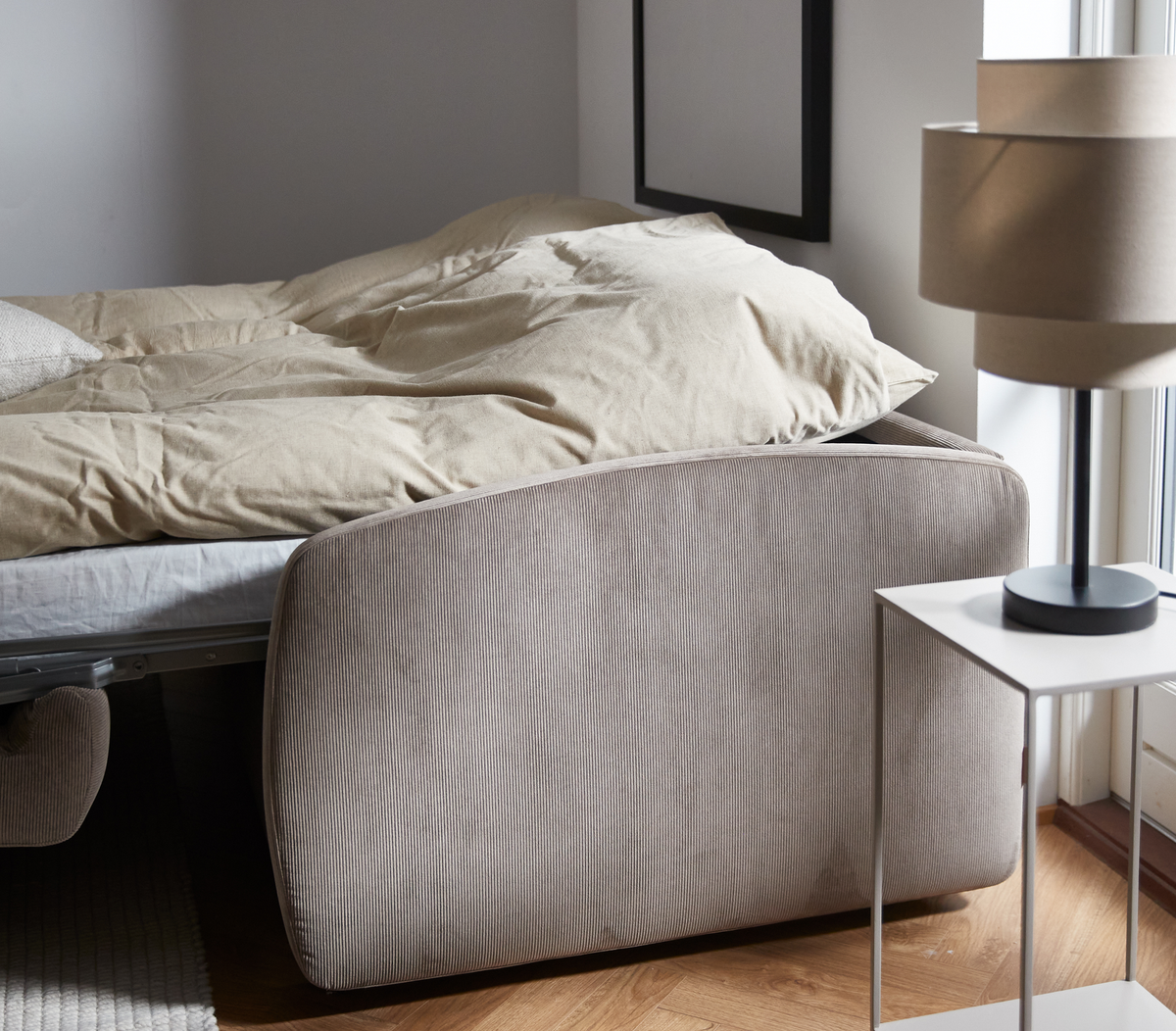 Säng med beige täcke intill vitt sängbord med beige bordslampa vid ett fönster; ljust trägolv och neutral inredning.