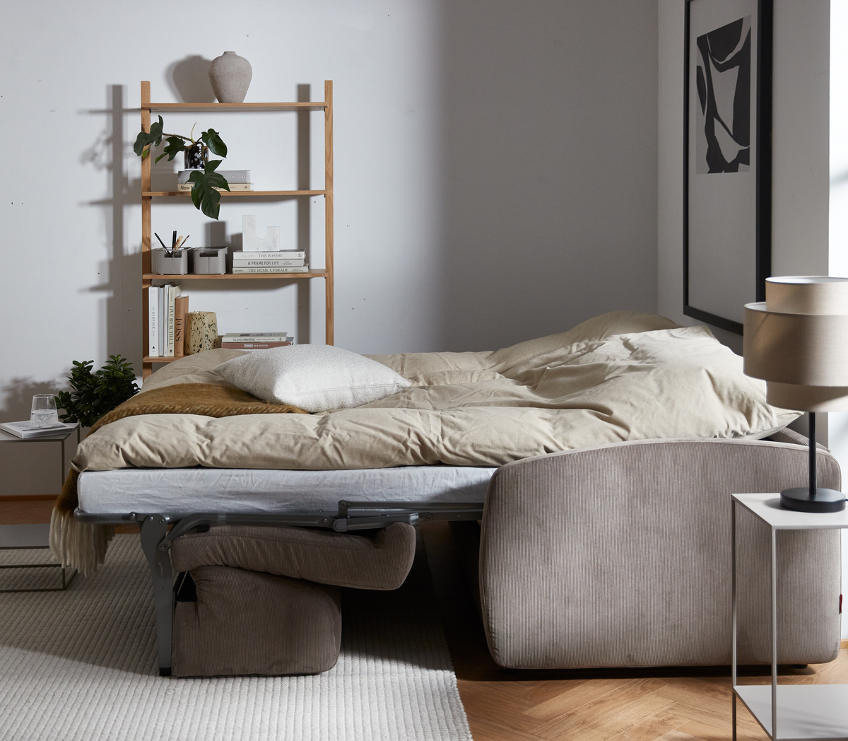 Uppbäddad bäddsoffa i beige med sidobord och bordslampa, bokhylla med böcker och växter i ett ljust vardagsrum.