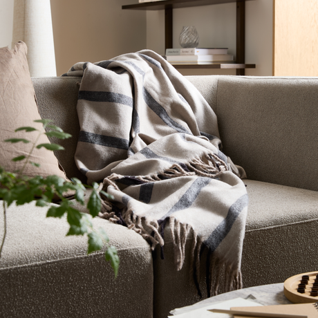 Beige soffa med mjuk, randig pläd med fransar; kudde, grön växt och hylla i bakgrunden i ett lugnt vardagsrum i dagsljus.