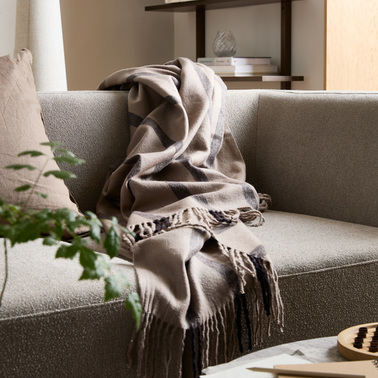 Beige rutmönstrad pläd med fransar hänger över en ljusgrå soffa i ett skandinaviskt vardagsrum.