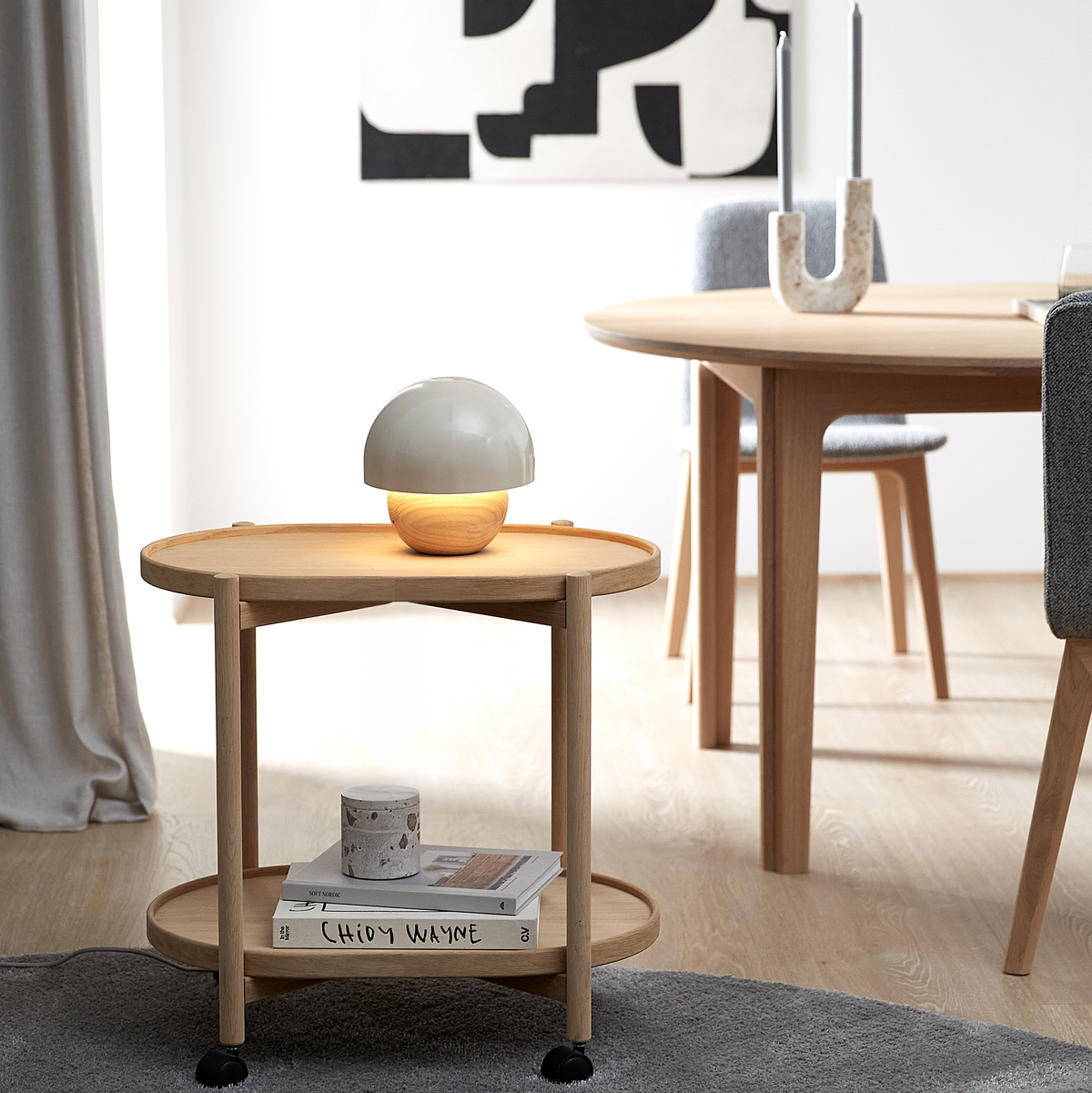 Ljust skandinaviskt rum med runt sidobord i trä och tänd kupollampa, runt matbord och grå stol bakom, abstrakt tavla och gardin; på underhyllan ligger böcker inklusive Chidy Wayne samt ett ljus.