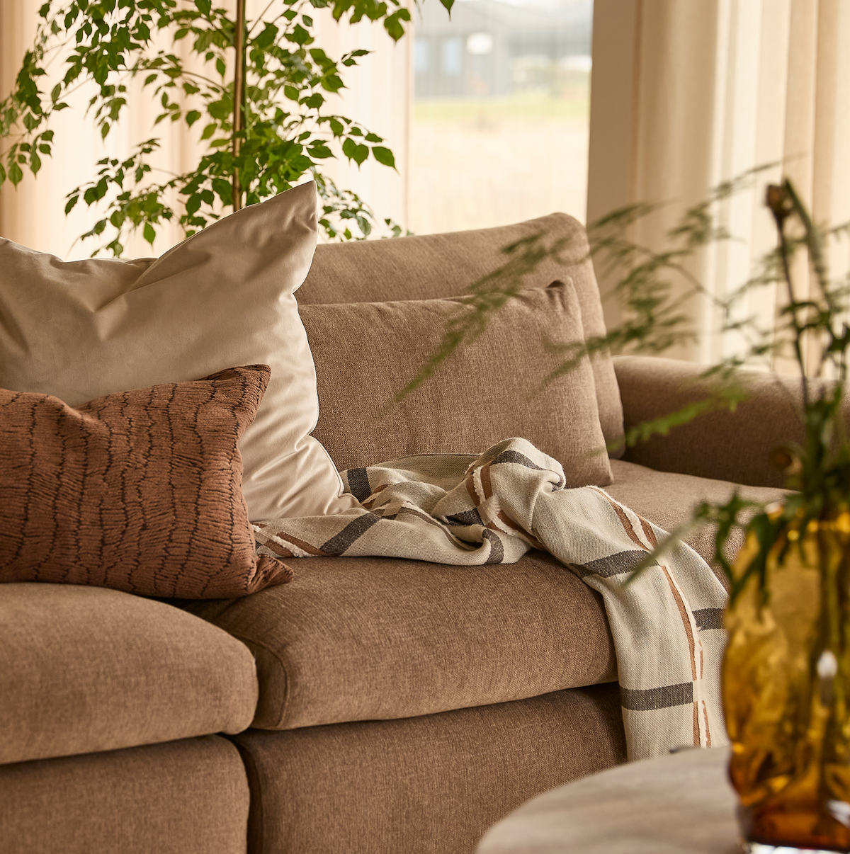 Närbild av en beige soffa med neutrala kuddar, en randig pläd över armstödet, gröna växter och en bärnstensfärgad vas i mjukt dagsljus.