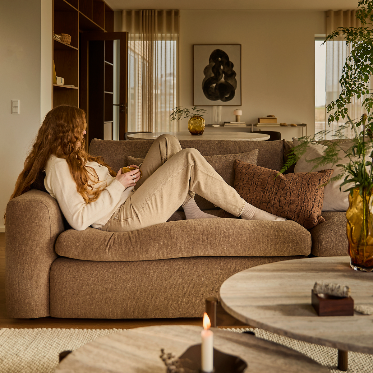 Person som ligger avslappnat på en beige soffa i ett ljust vardagsrum med kuddar, växter, ljus och konst på väggen.