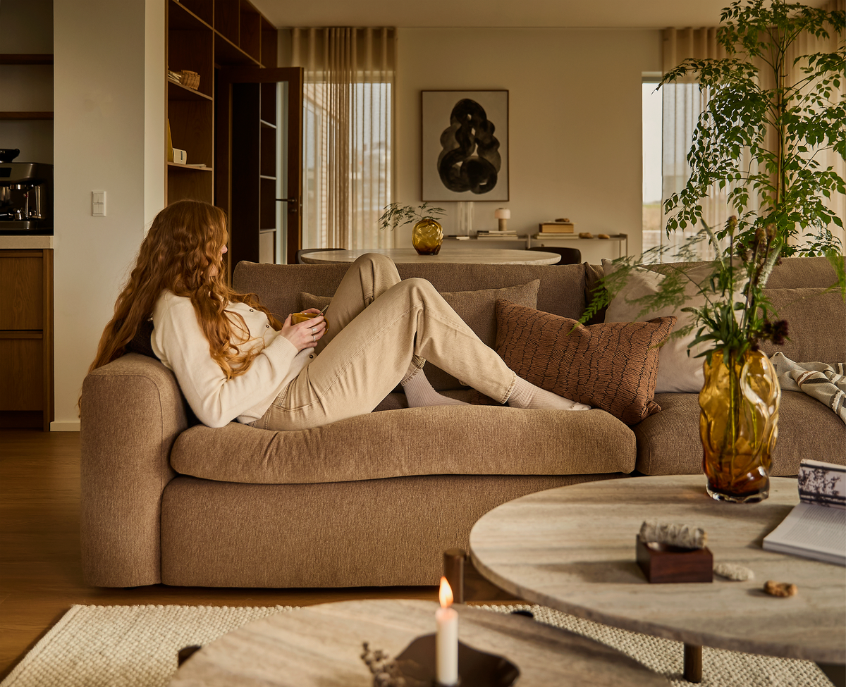 Person ligger avslappnat på en beige soffa i ett varmt, skandinaviskt vardagsrum med gröna kvistar i bärnstensfärgad vas, tänd ljus och runda soffbord.