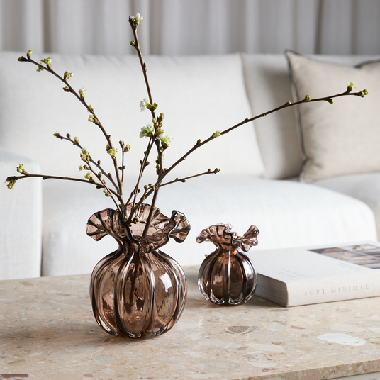 Brun rökfärgad glasvas med veckad kant och blommande kvistar på ett soffbord i sten, bredvid en mindre vas och en bok, Soft Minimal, med ljus soffa i bakgrunden.