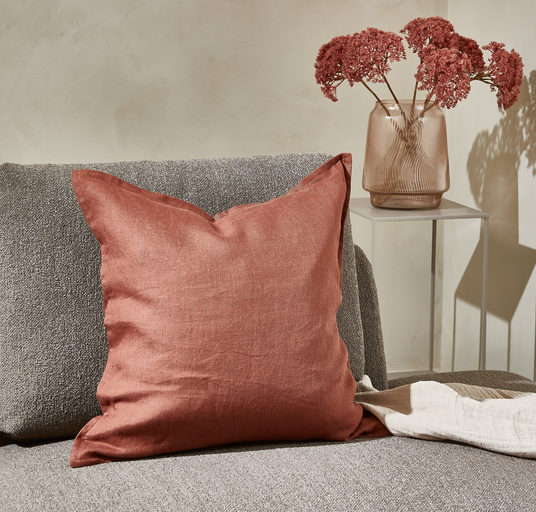 Rostfärgad linneskudde på grå soffa bredvid glasvas med torkade blommor.