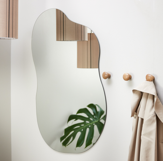 Organisk väggspegel på vit vägg med runda träknoppar, beige kappa och en monstera som speglas; pendellampa syns i reflektionen.