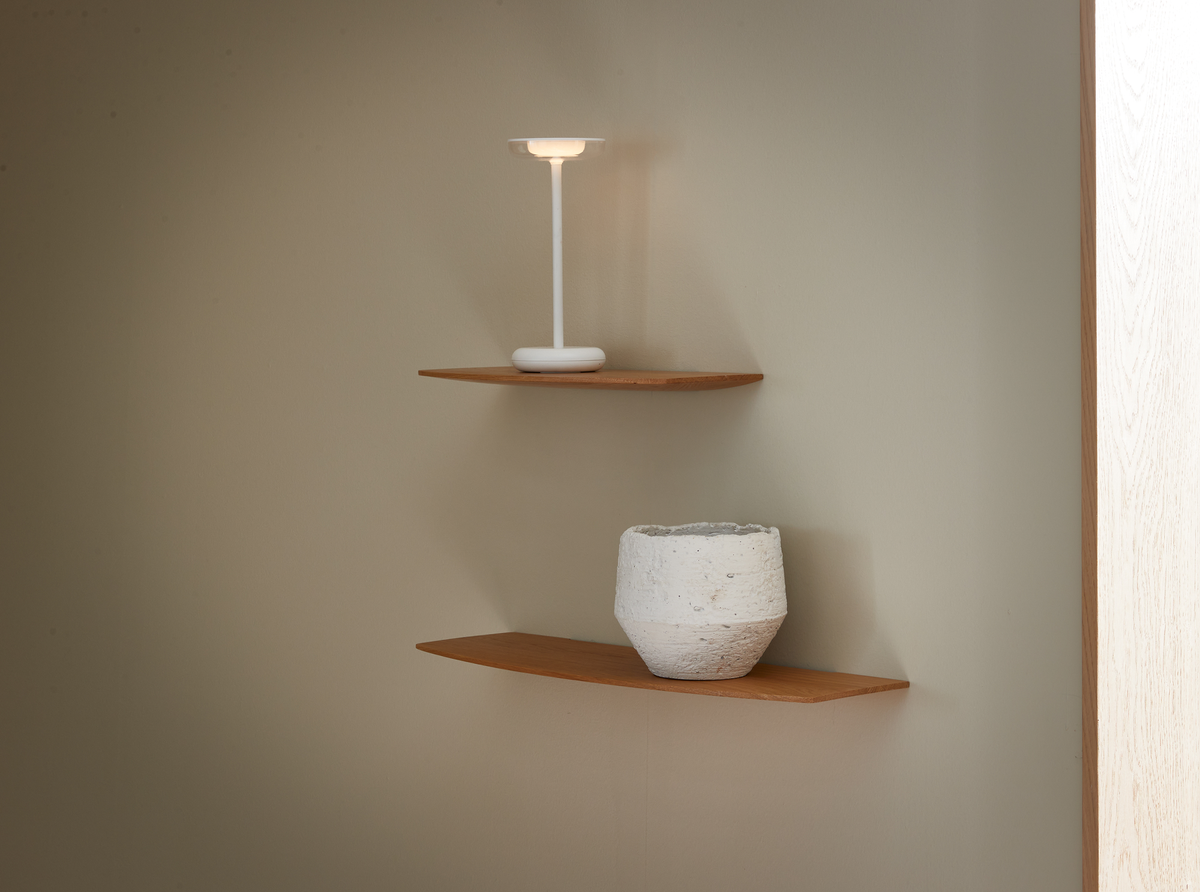 Två vägghyllor i trä med en vit bärbar bordslampa på den övre och en vit strukturerad vas på den nedre, mot en beige vägg.
