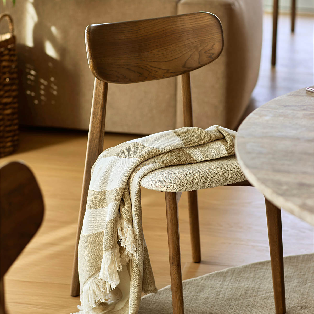 Trästol vid rund bordsskiva med beige‑vit randig pläd med fransar över sitsen i ett ljust, varmt matplats/vardagsrum.