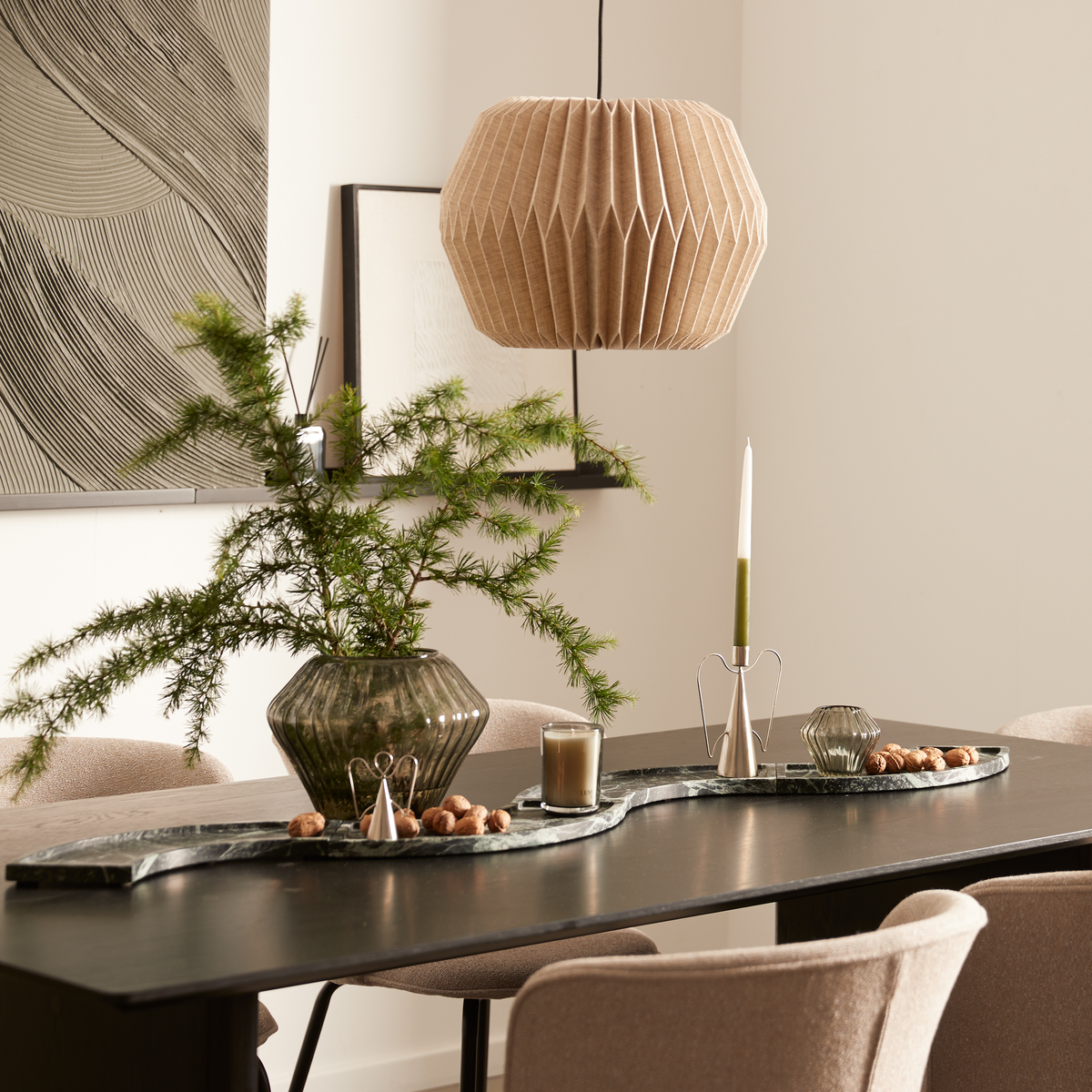 Stilat matbord med grön glasvas med granris, skulptural ljusstake med grönt kronljus och litet ljus på en stenbricka; veckad pendellampa och abstrakt väggkonst i en lugn, neutral matsal.