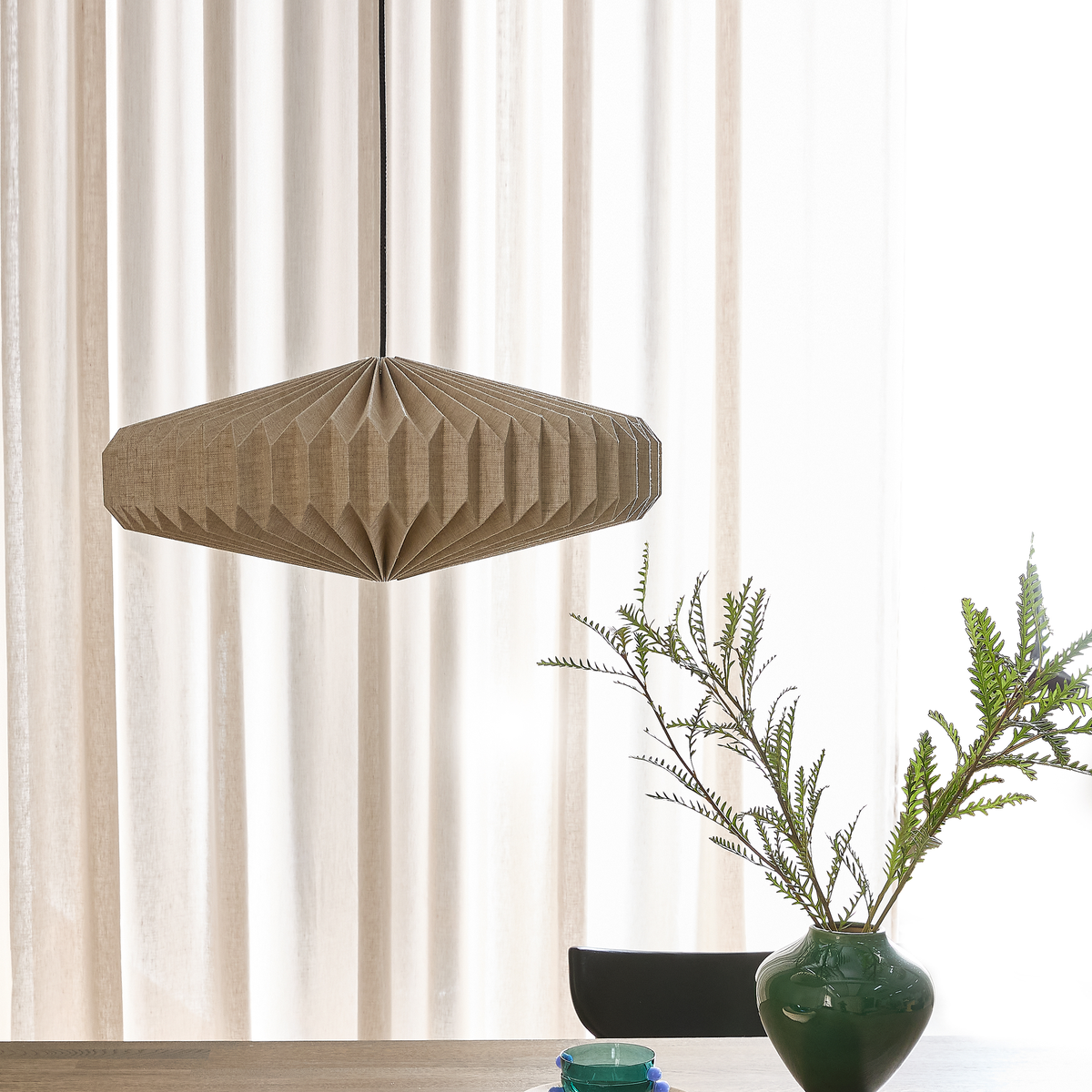 Vikmönstrad beige pendellampa över ett bord med grön vas och kvistar framför ljusa gardiner.