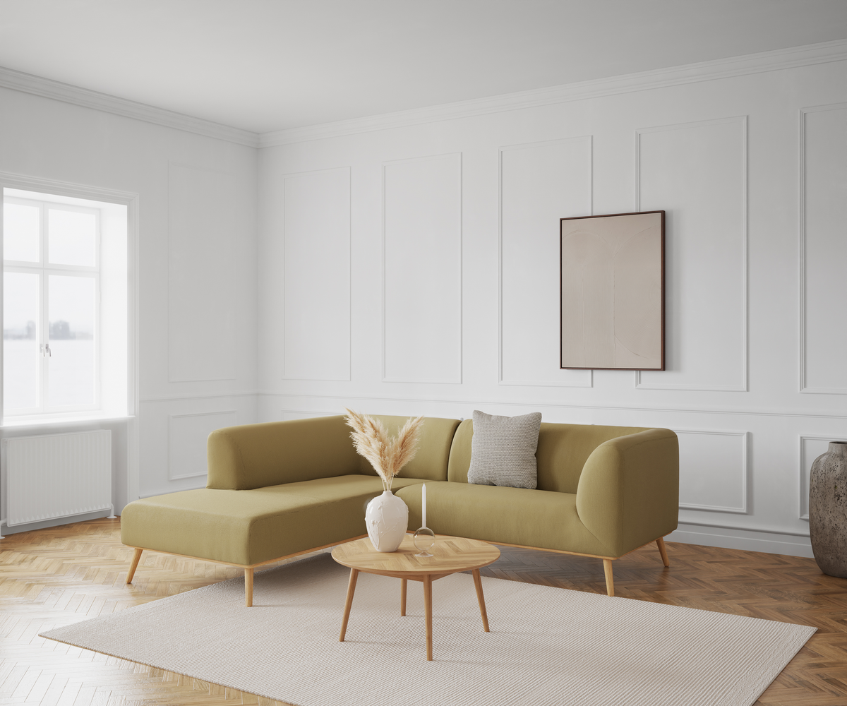 Ljus vardagsrumsinteriör med olivgrön hörnsoffa, ljust träbord med vas med pampasgräs, beige matta och enkel väggtavla mot vit panelvägg vid fönster.