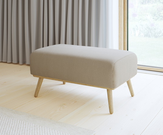 Beige stoppad bänk/pall med träben på ljust trägolv, framför grå gardiner och ett fönster i dagsljus.