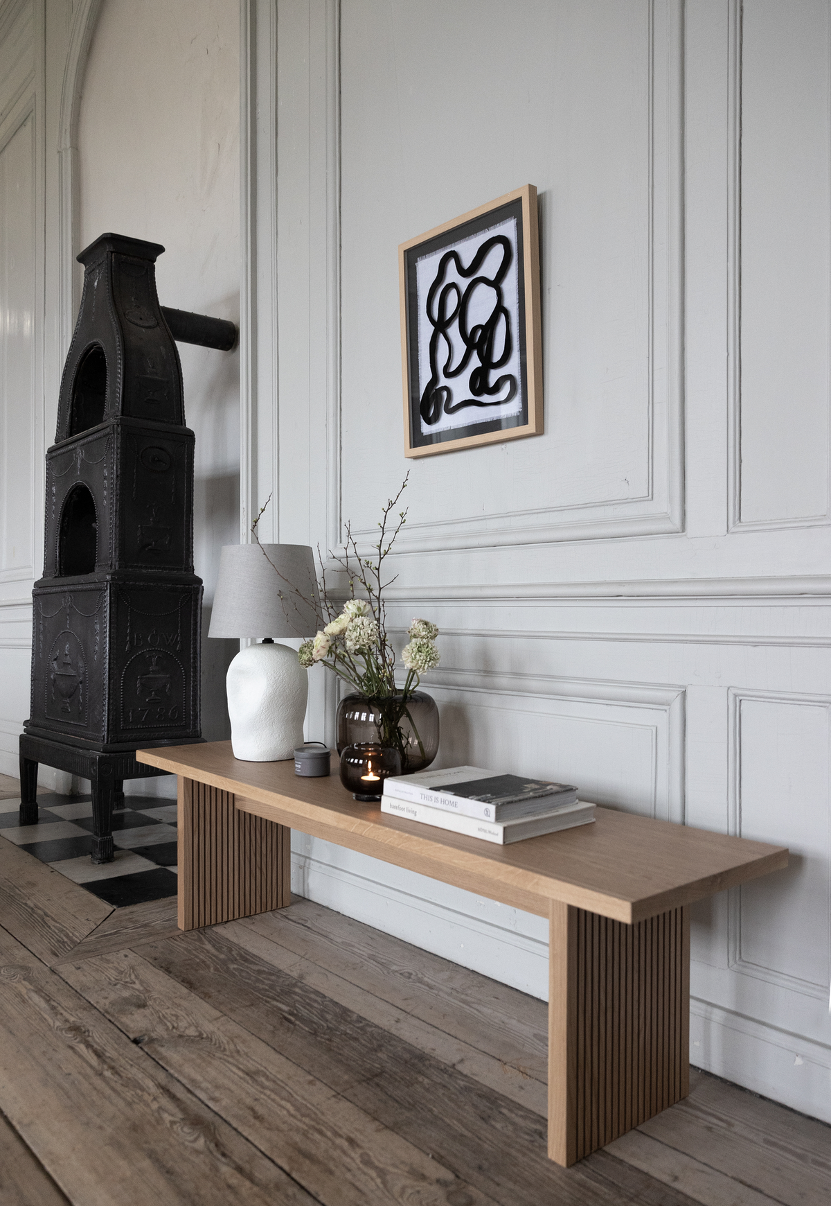 Ljus skandinavisk interiör med en träbänk mot en panelvägg, bordslampa, vas med blommor, mörk glasvas och staplade böckerna ”This Is Home”; en svart gjutjärnsspis och en abstrakt tavla på väggen.