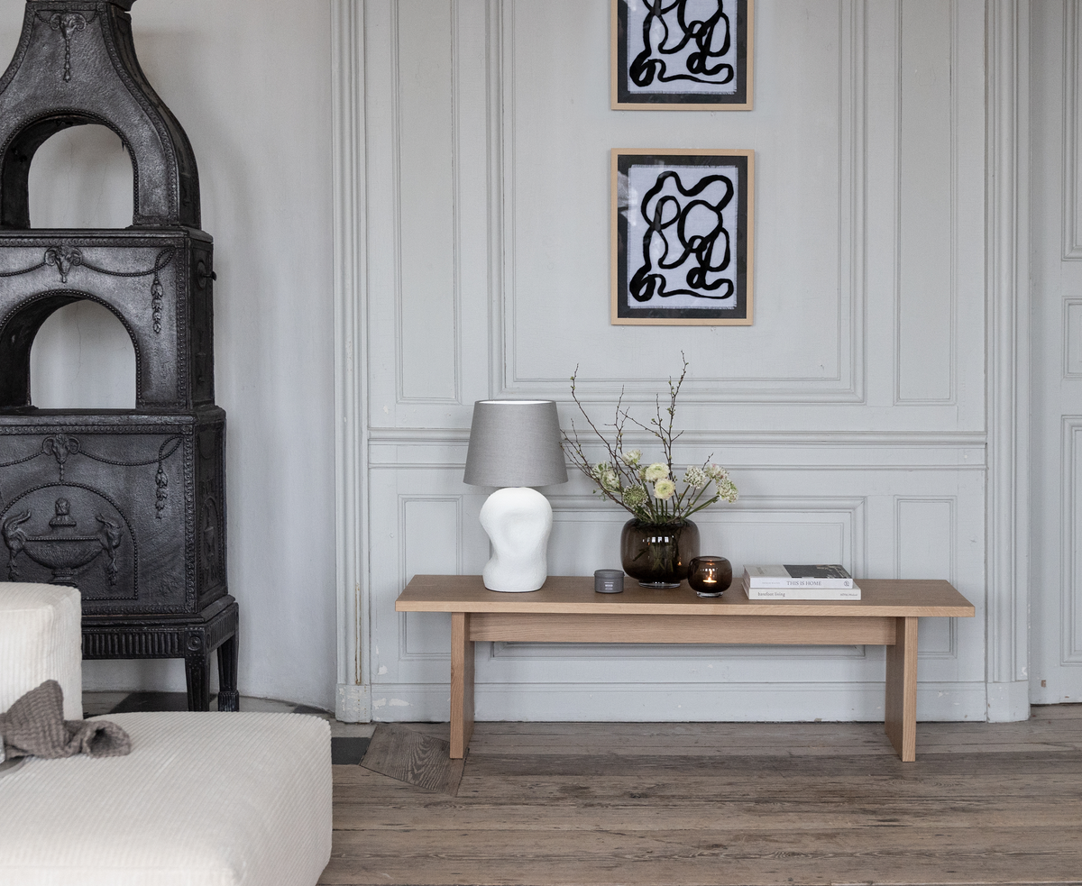 Ljusgrå panelvägg med träbänk, bordslampa, glasvas med grenar och blommor, ljus och böcker; två abstrakta svartvita tavlor ovanför samt gjutjärnskamin och beige soffa i en lugn skandinavisk vardagsrumsmiljö.