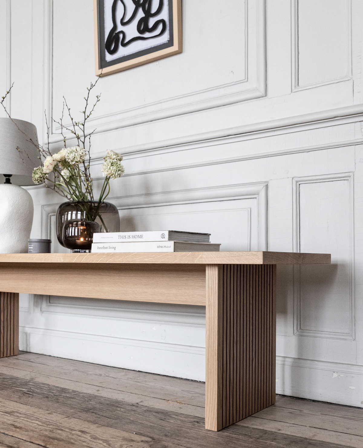 Ljus träbänk mot vit panelvägg med abstrakt tavla ovanför; på bänken står en brun glasvas med vita blommor, en vit bordslampa och två böcker med titlarna ”THIS IS HOME” och ”barefoot living” – lugn, minimalistisk skandinavisk inredning.