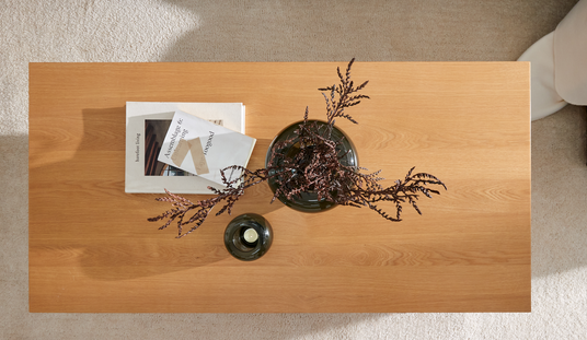 Ovanifrån vy av ett rektangulärt soffbord i ljust trä med två böcker (”barefoot living” och ”Assemblage 6; toogood”), en mörk glasvas med torkade kvistar och en liten ljushållare på en ljus matta.
