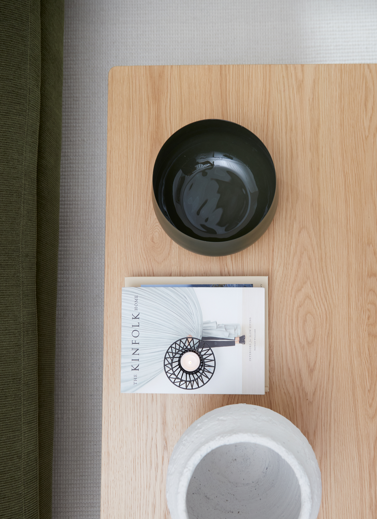 Ljus träbord med en mörk skål, ett värmeljus i svart ljushållare ovanpå boken THE KINFOLK HOME och en ljus betongvas; grön soffa i kanten.