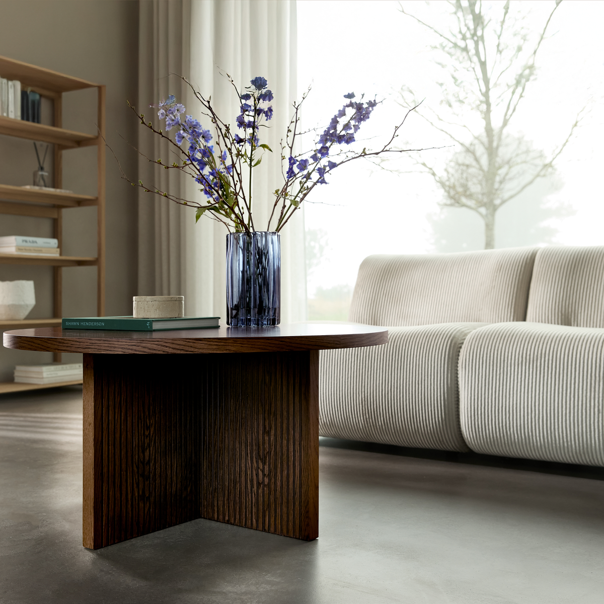 Mörkt soffbord i trä med blå glasvas med lila blommor och en bok, framför en ljus ribbad soffa vid ett stort fönster med skira gardiner.