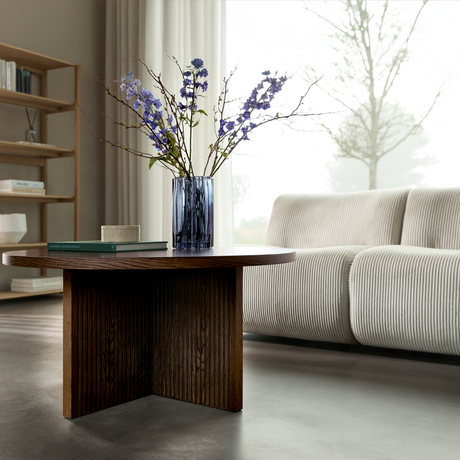 Mörkt soffbord i trä med blå glasvas med lila blommor och en bok, framför en ljus ribbad soffa vid ett stort fönster med skira gardiner.
